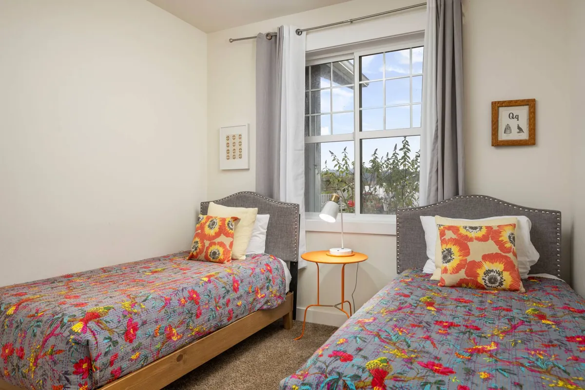 A cheerful bedroom characterized by colorful bedspreads on twin beds, decorative pillows, a nightstand with a lamp, and a window with greenery outside. The playful yet calming palette is perfect for families or children.