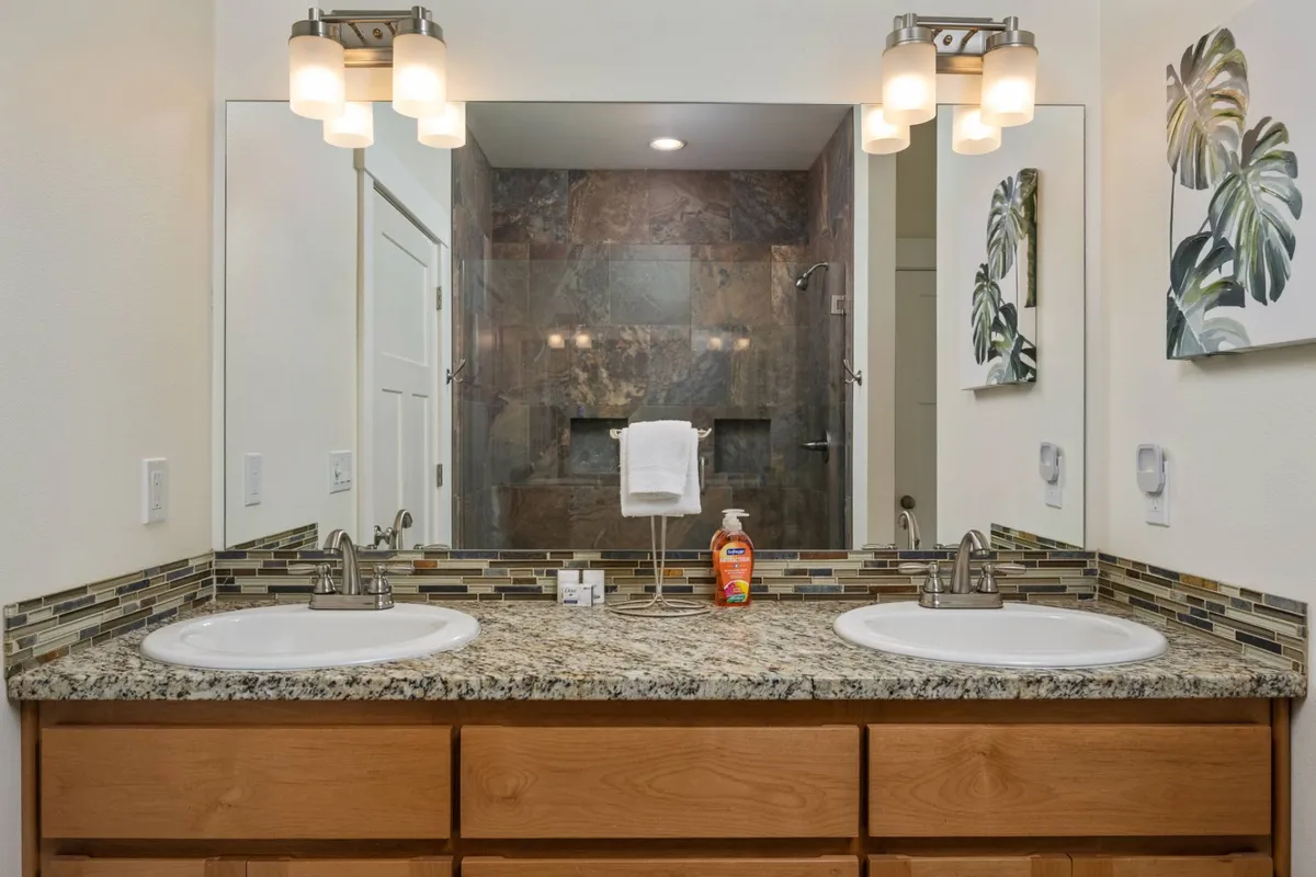 A bathroom scene with double vanity sinks, granite countertops, a large framed mirror, and decorative artwork. The room is fresh, spacious, and ideal for morning or evening routines.