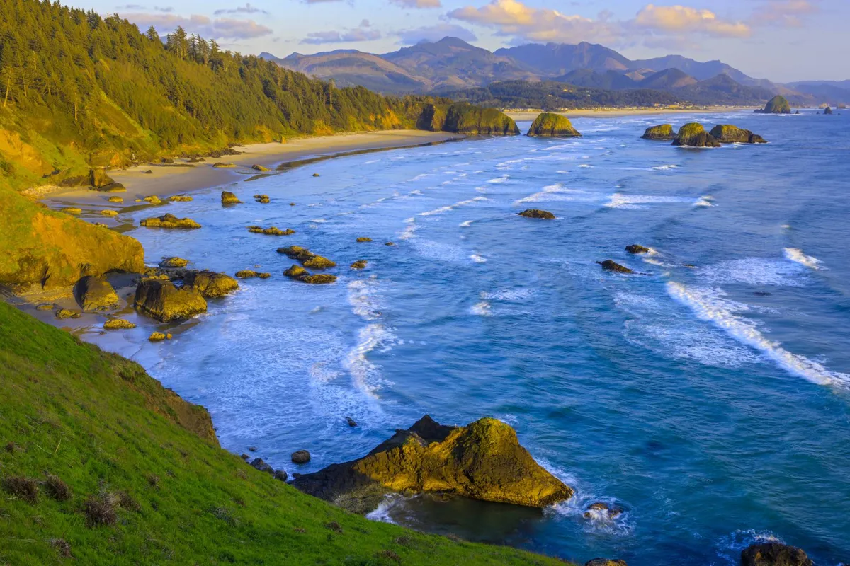 A sweeping view of the rocky shoreline, with gentle waves lapping against the coast and rolling hills in the background. Distant mountains are visible on the horizon, capturing the rugged natural beauty and sense of adventure the Oregon Coast offers.