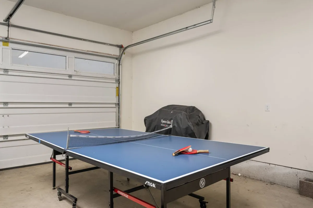 A bright, energetic indoor game space featuring a full-size ping pong table, paddles ready for play, and a large garage door that hints at strong indoor-outdoor connectivity. The setting is ideal for family tournaments and friendly competition.