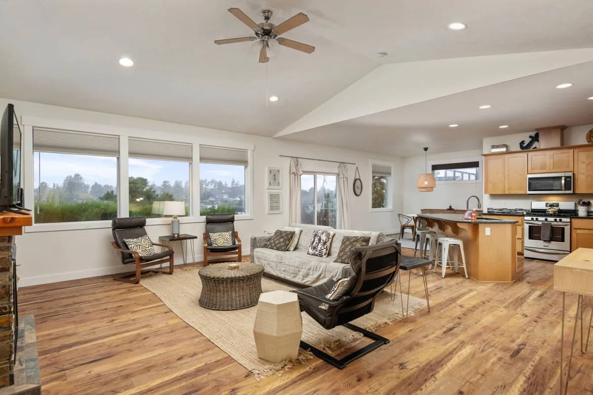 The image showcases a bright, welcoming living area with a cozy sofa adorned with decorative pillows, a stylish wooden coffee table, and a modern kitchen island with bar stools. Sunlight pours in through expansive windows, creating a warm, inviting atmosphere perfect for gathering with family or friends. The open layout encourages relaxation, conversation, and shared meals, all while offering glimpses of the tranquil outdoors.