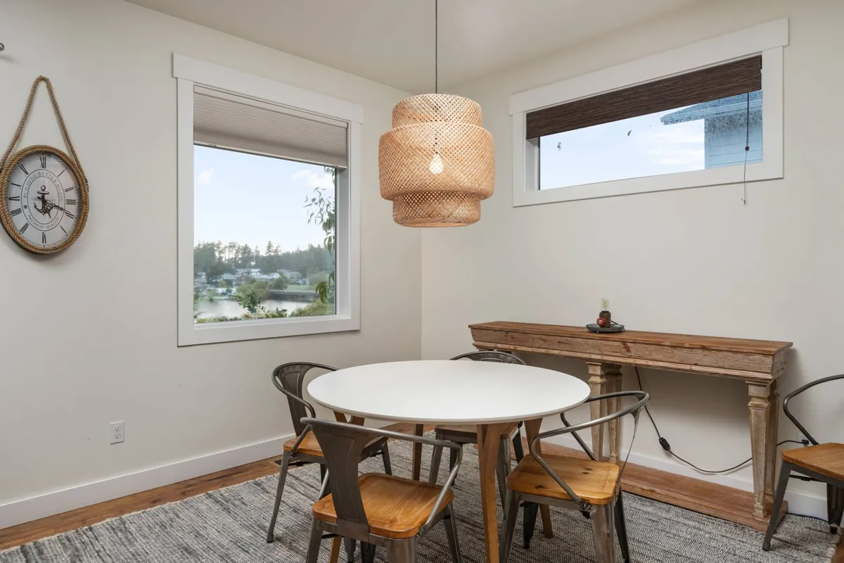 The image shows a dining area with a modern table set beneath a striking hanging light fixture, with a large window offering a glimpse of the landscape outside. The setting looks welcoming and perfect for guests to gather, share meals, or watch the sunset together.
