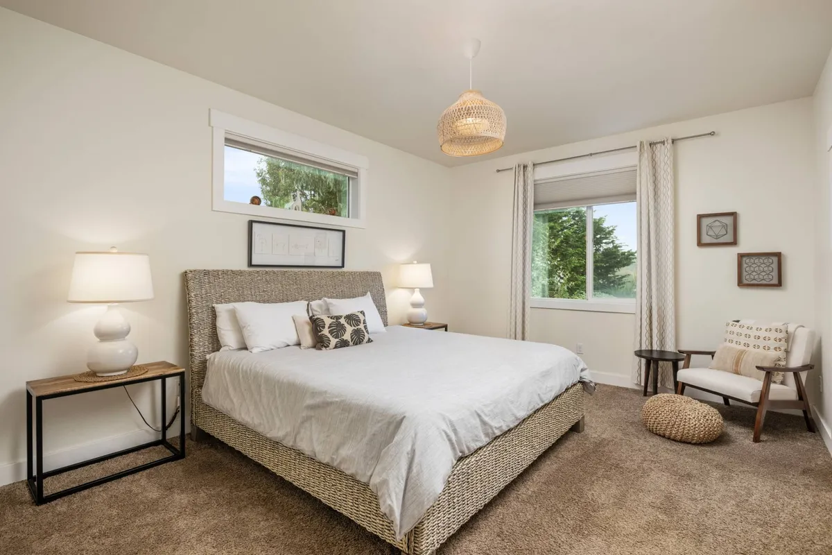 A bright and inviting bedroom featuring a neatly made bed with crisp linens, large windows letting in natural light, contemporary light fixtures, and an accent chair perfect for relaxation or reading. Soft décor and a tranquil atmosphere create an ideal sleeping retreat.