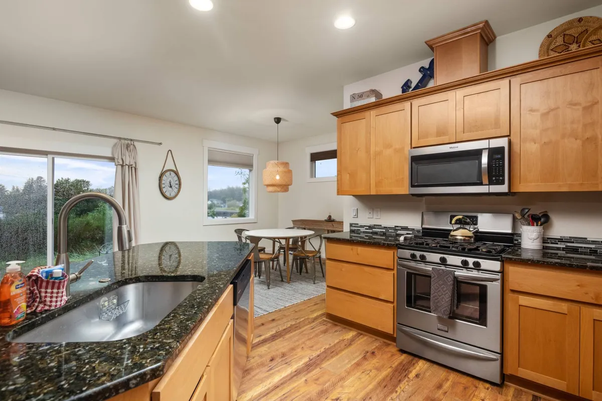An open-concept dining and kitchen area featuring a granite countertop, stainless steel appliances, wooden cabinetry, and a dining table set by large windows. The space is designed for social gatherings, family meals, and entertaining friends.